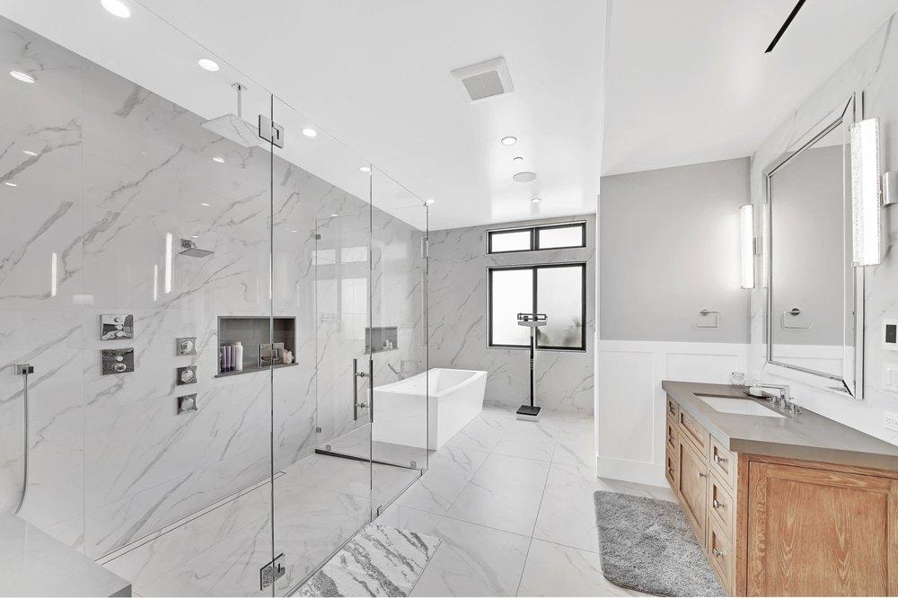 Bathroom remodel in Newport Beach CA — full marble slab walk-in shower with frameless glass enclosure, freestanding soaking tub, wood vanity with quartz countertop, and large format marble tile flooring by Main Street Kitchen & Flooring