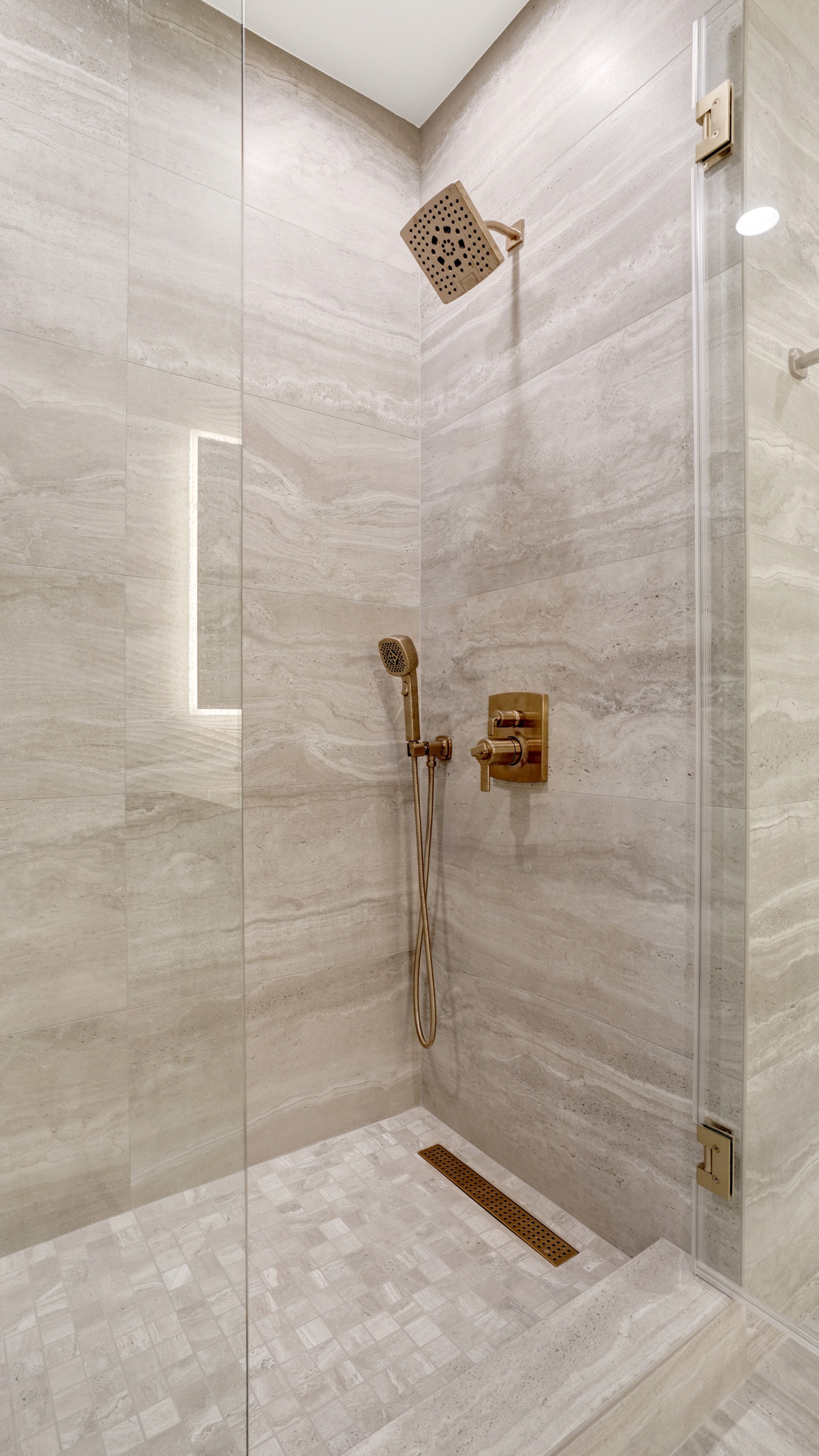 Bathroom remodel in Santa Ana CA — frameless glass shower with floor-to-ceiling travertine-look tile, brass rain showerhead, handheld wand, thermostatic valve, and linear drain by Main Street Kitchen & Flooring