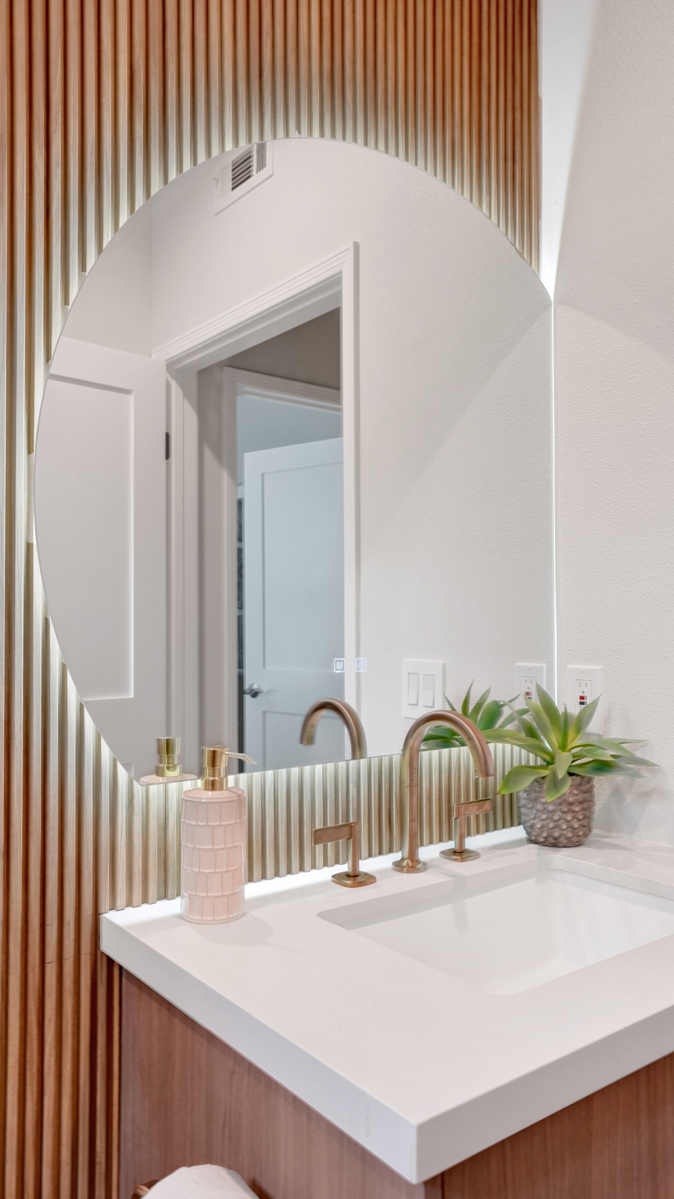 Bathroom remodel in Santa Ana CA — closeup of floating vanity with round frameless mirror, vertical wood slat accent wall, fluted glass tile backsplash, brass widespread faucet, and succulent decor by Main Street Kitchen & Flooring