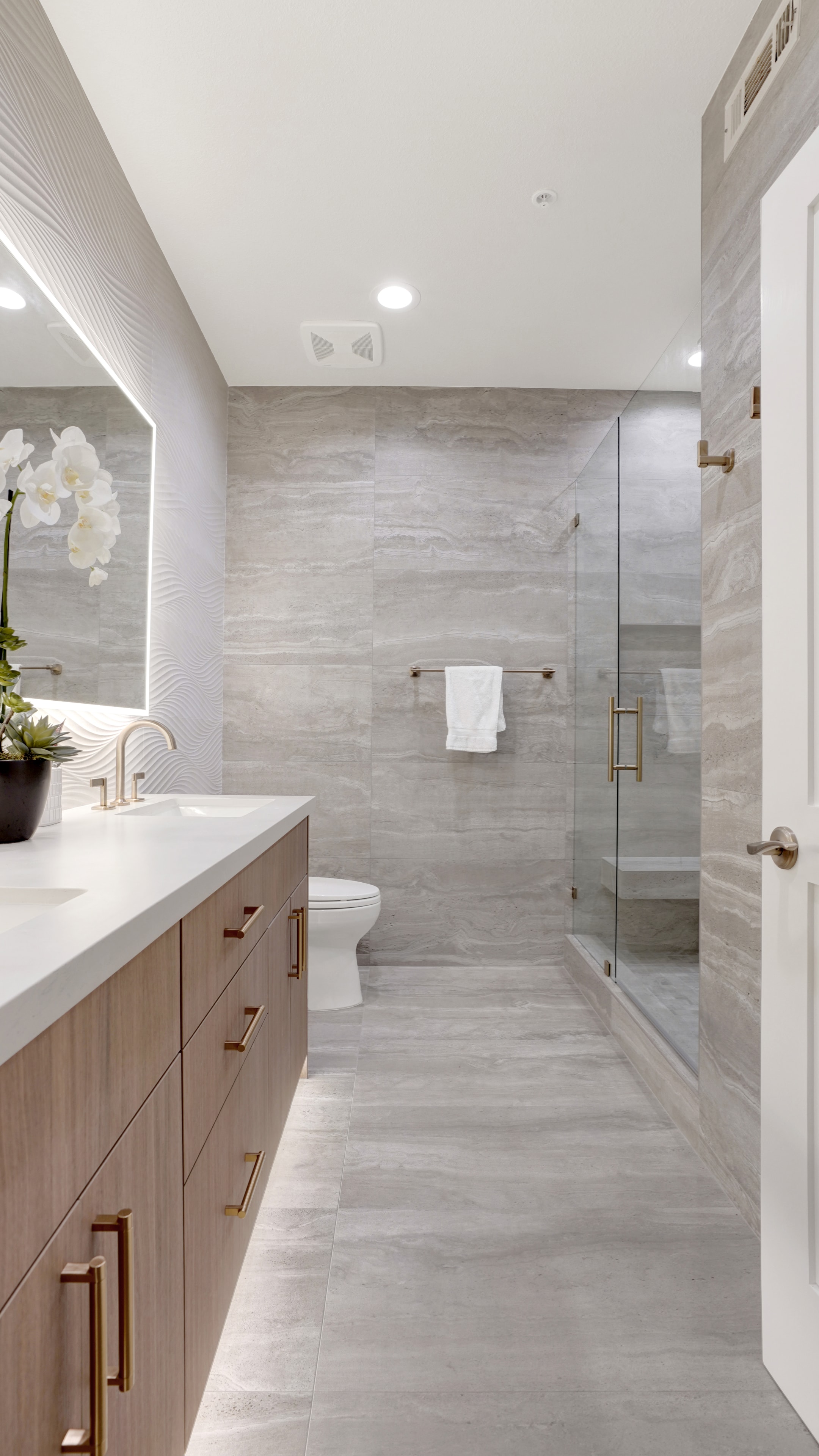 Bathroom remodel in Santa Ana CA — wood floating vanity, travertine-look large format tile walls and floor, frameless glass shower enclosure with brass hardware, and backlit LED mirror by Main Street Kitchen & Flooring