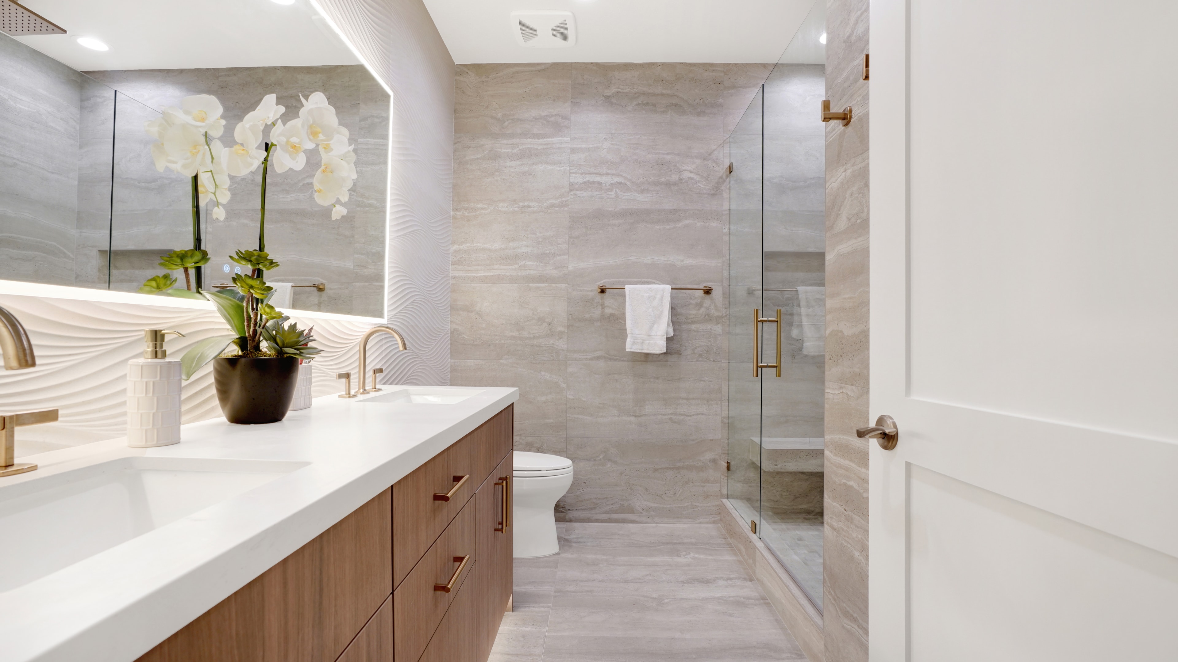 Bathroom remodel in Santa Ana CA — wood floating vanity with white quartz countertop, 3D wave tile backsplash, brass fixtures, backlit LED mirror, and frameless glass shower with travertine-look tile by Main Street Kitchen & Flooring