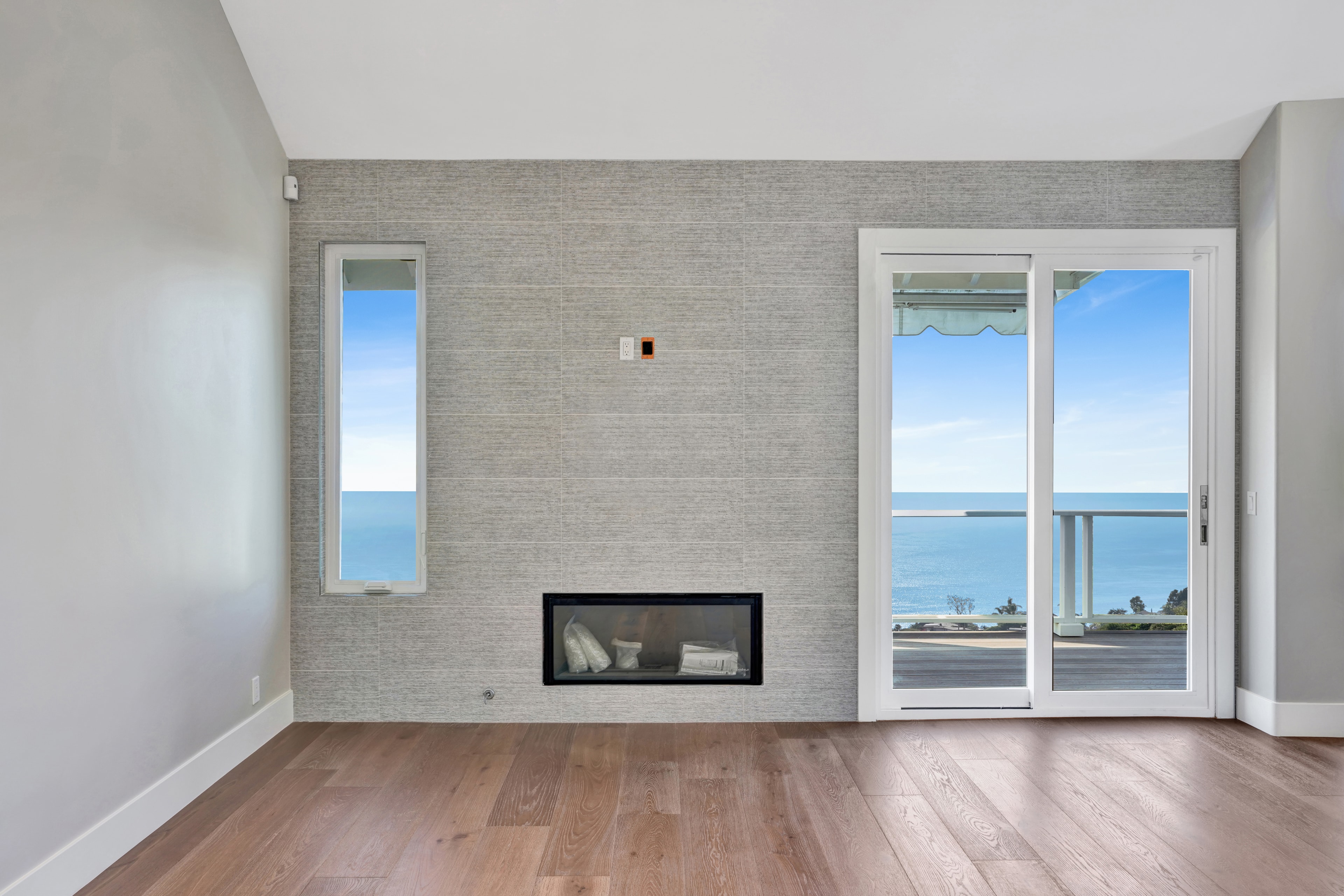 Home remodel in Orange County CA — bright living room with large-format linen-texture tile fireplace feature wall, linear gas fireplace, wide-plank hardwood flooring, and floor-to-ceiling sliding glass doors with panoramic ocean view by Main Street Kitchen & Flooring