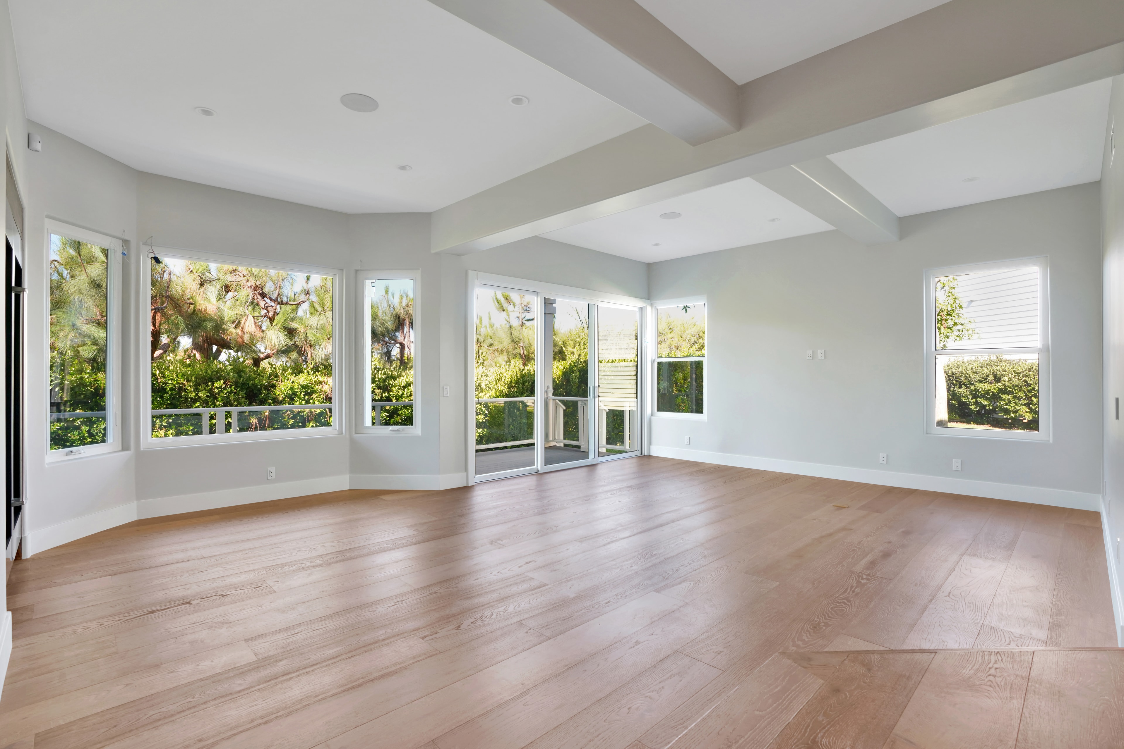 Home remodel in Orange County CA — open room with wide-plank hardwood flooring, coffered ceiling with exposed beams, curved bay windows, and French doors overlooking lush garden by Main Street Kitchen & Flooring