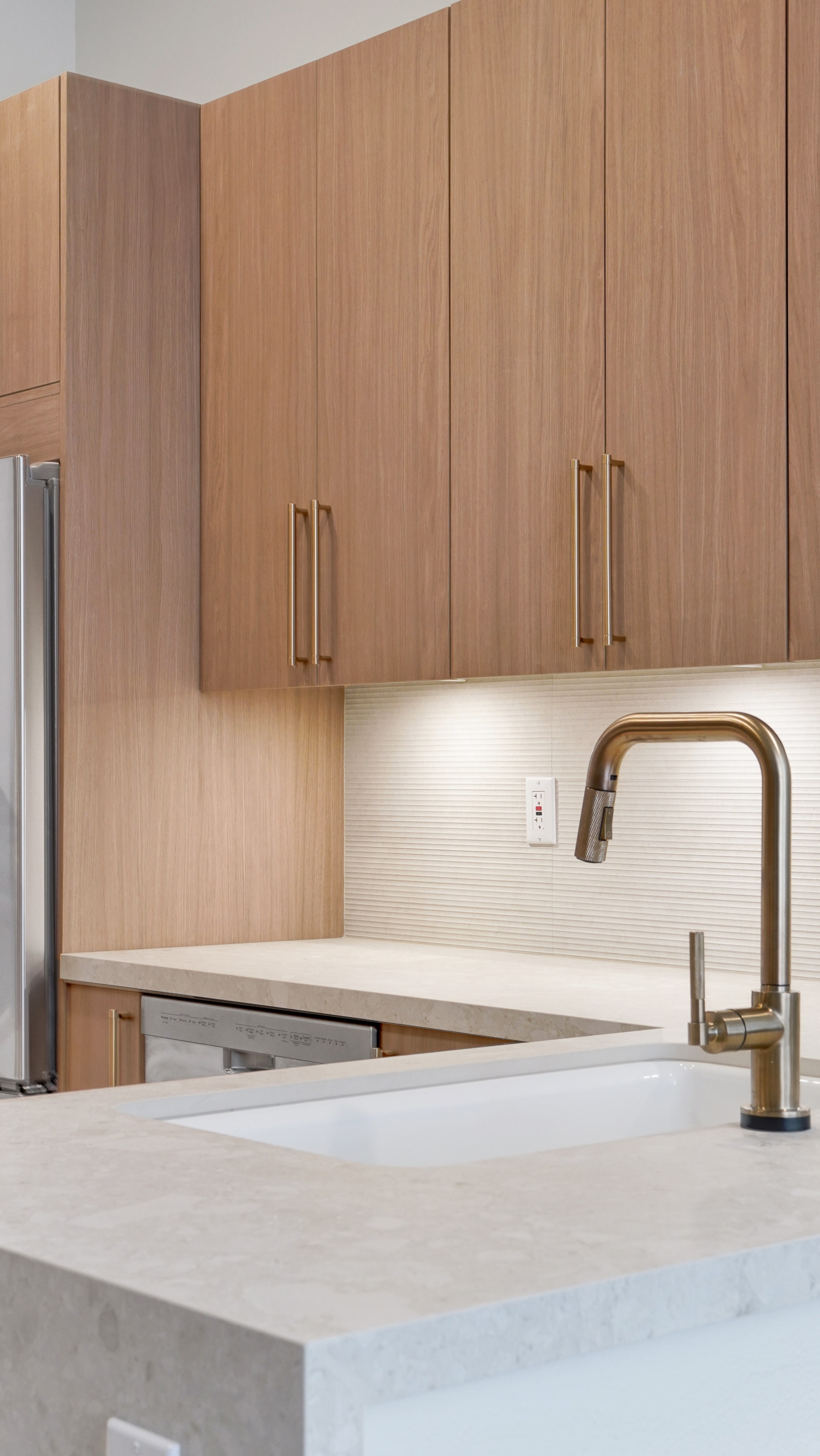 Kitchen remodel in Irvine CA — closeup of warm wood flat-panel cabinets with brass bar pulls, fluted ceramic tile backsplash with under-cabinet lighting, white quartz countertop, and brass pull-down kitchen faucet by Main Street Kitchen & Flooring