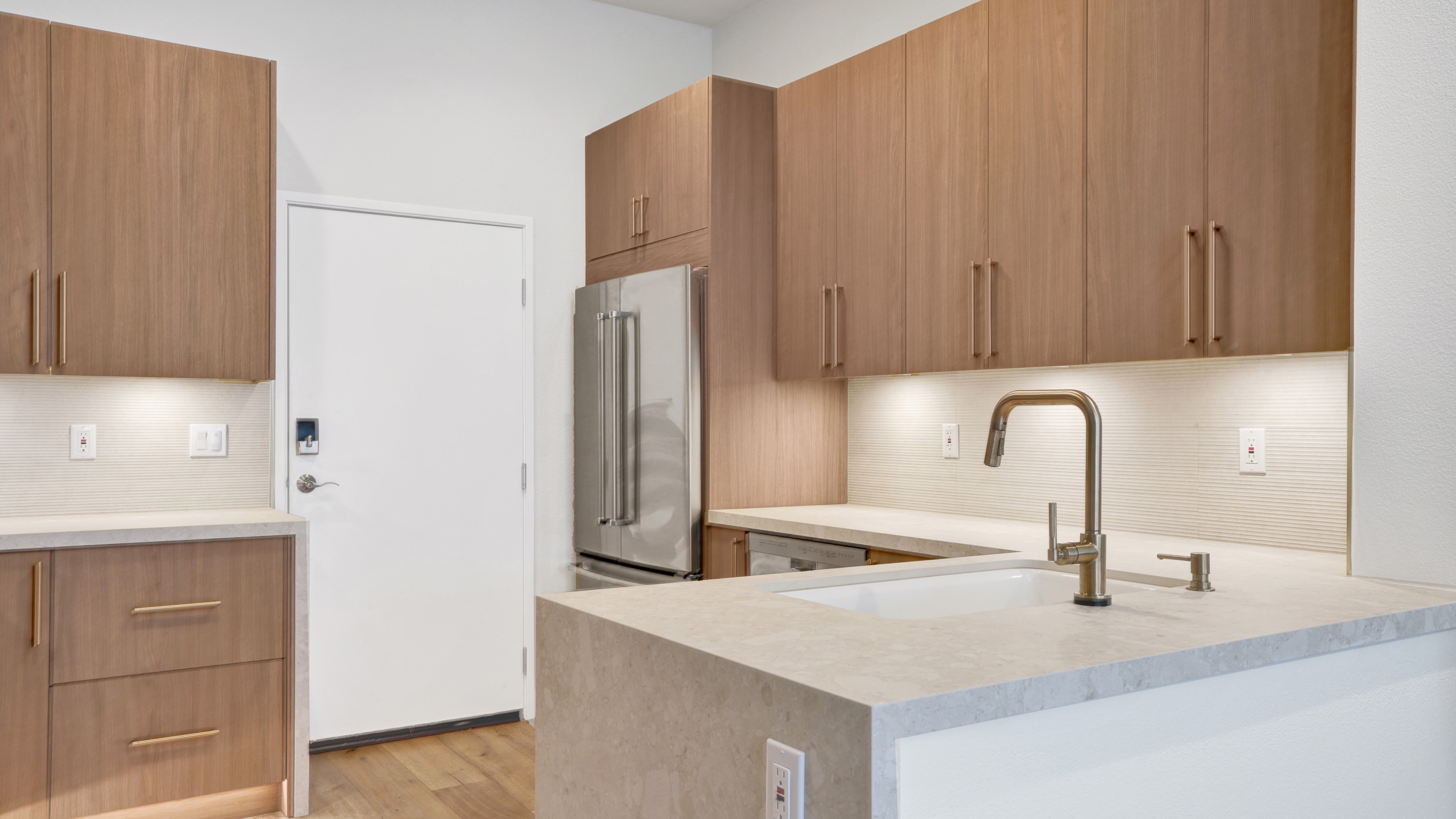 Kitchen remodel in Irvine CA — wood flat-panel upper and lower cabinets with brass hardware, white quartz countertop, undermount sink with brass pull-down faucet, fluted ceramic tile backsplash, and stainless refrigerator by Main Street Kitchen & Flooring