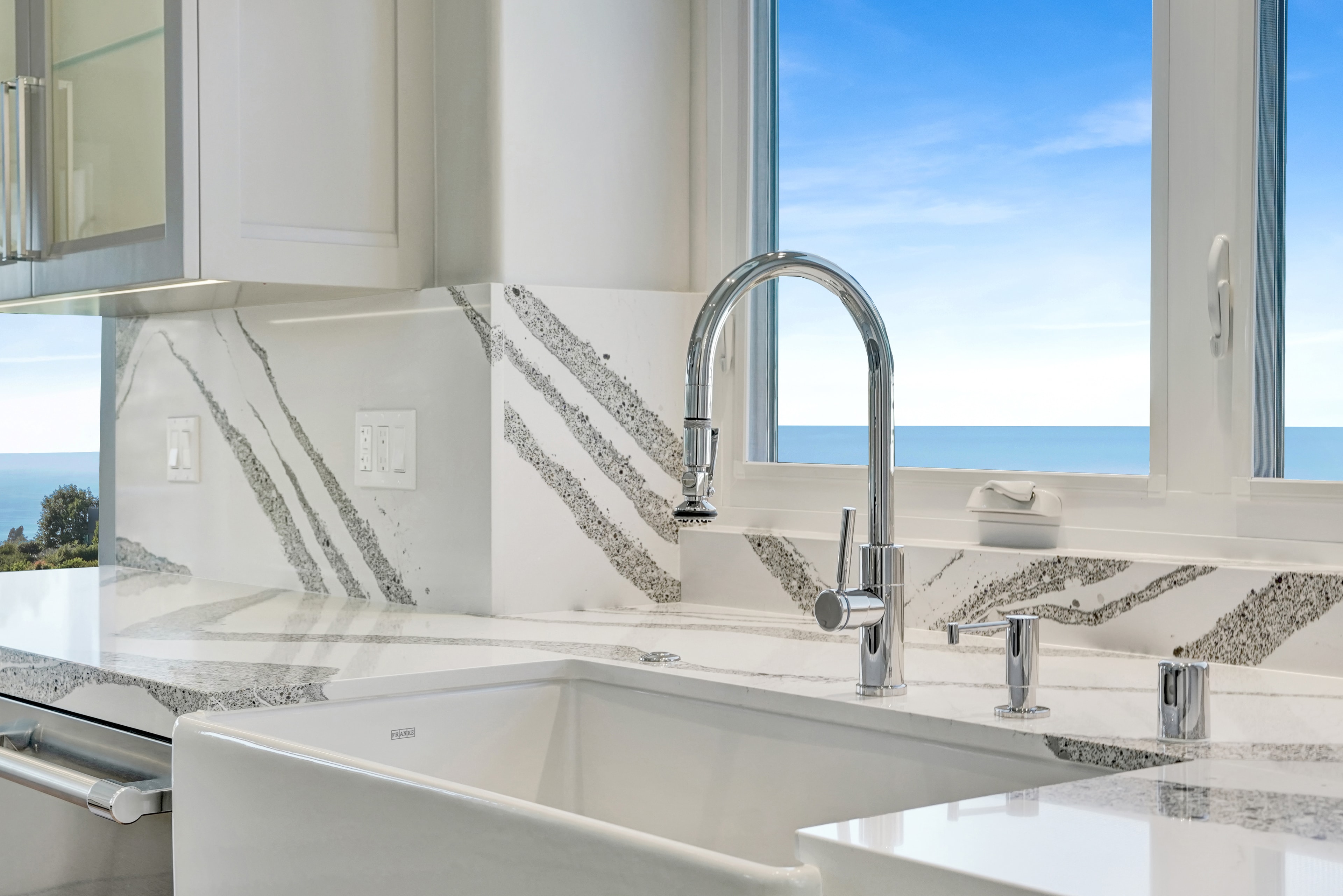 Kitchen remodel in Laguna Beach CA — white shaker cabinets with farmhouse apron sink, chrome pull-down faucet, book-matched quartz backsplash, stainless dishwasher, and direct ocean view through window by Main Street Kitchen & Flooring