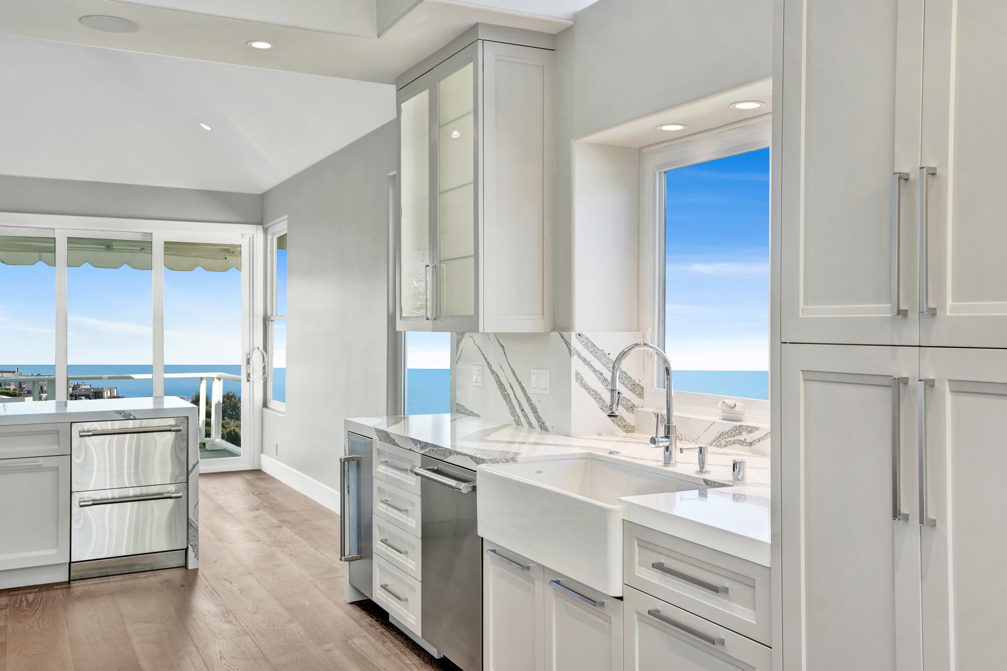 Kitchen remodel in Laguna Beach CA — galley view of white shaker cabinetry with full-height stainless refrigerator, professional range, farmhouse sink, light hardwood floor, and ocean view through sliding glass doors by Main Street Kitchen & Flooring
