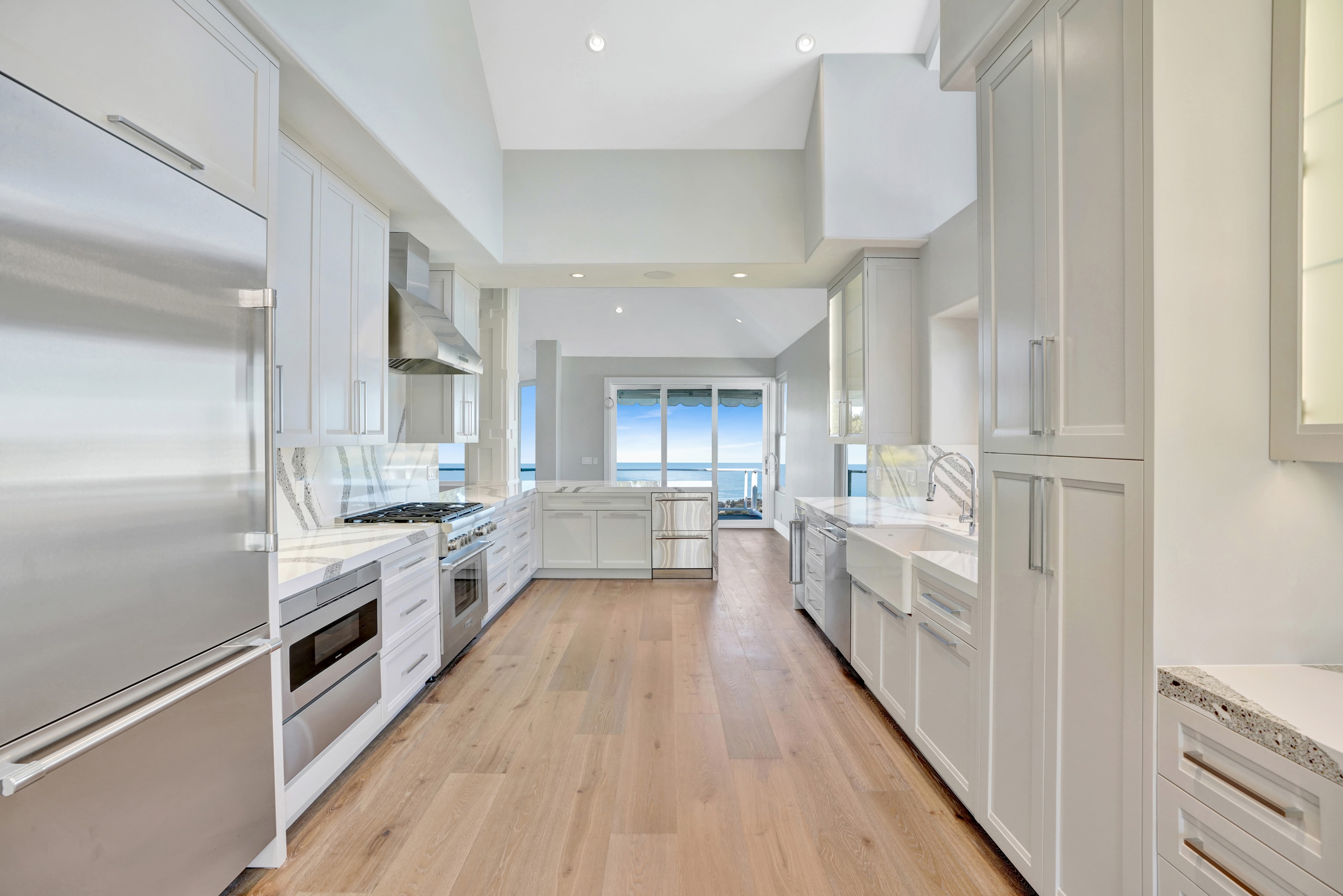 Kitchen remodel in Laguna Beach CA — closeup of white shaker cabinets with dramatic book-matched quartz slab backsplash, professional 6-burner gas range, stainless hood, and ocean view through window by Main Street Kitchen & Flooring