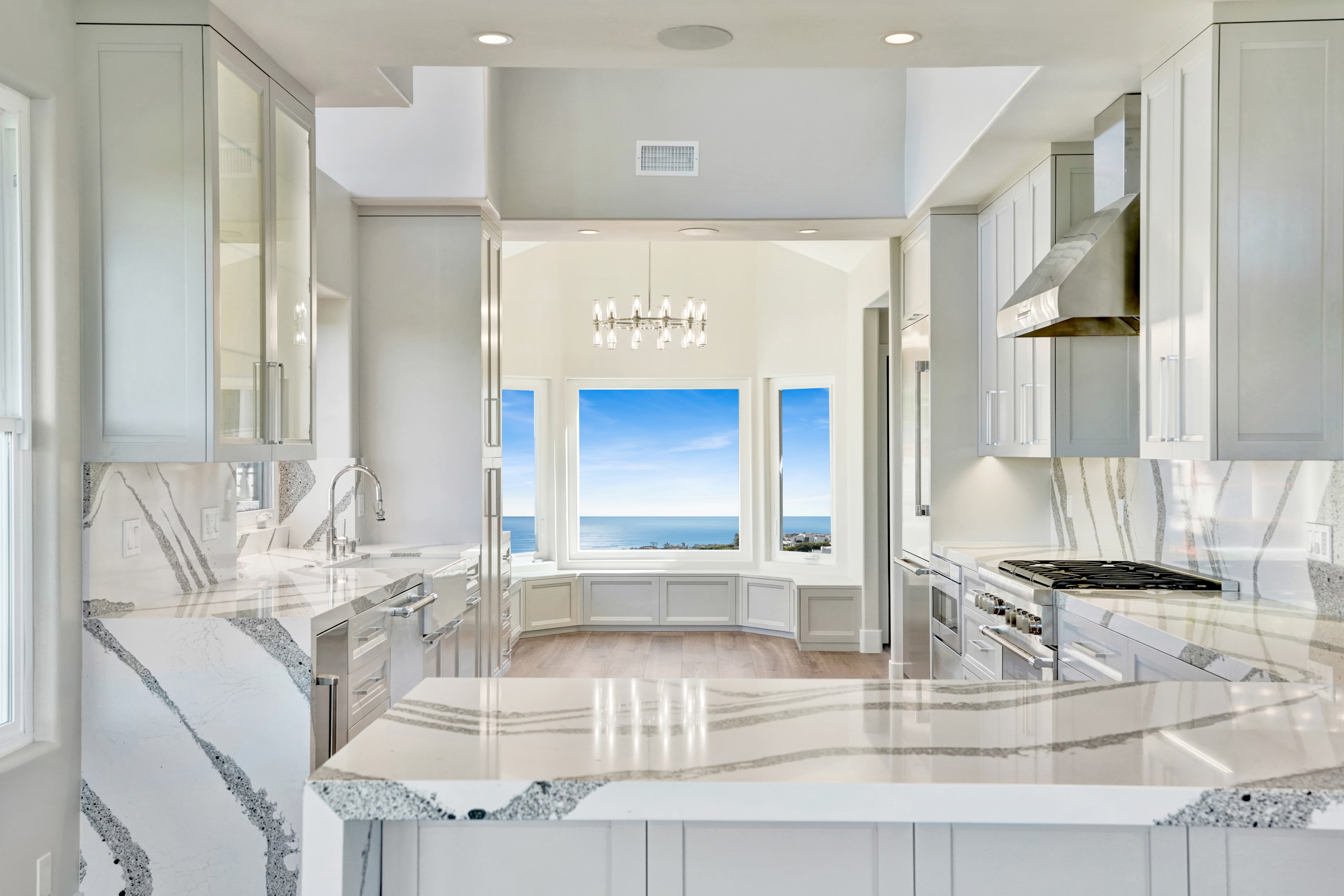Kitchen remodel in Laguna Beach CA — wide view of white shaker cabinetry with book-matched quartz waterfall island countertop and full-height backsplash, chandelier, stainless appliances, and panoramic ocean view through bay window by Main Street Kitchen & Flooring