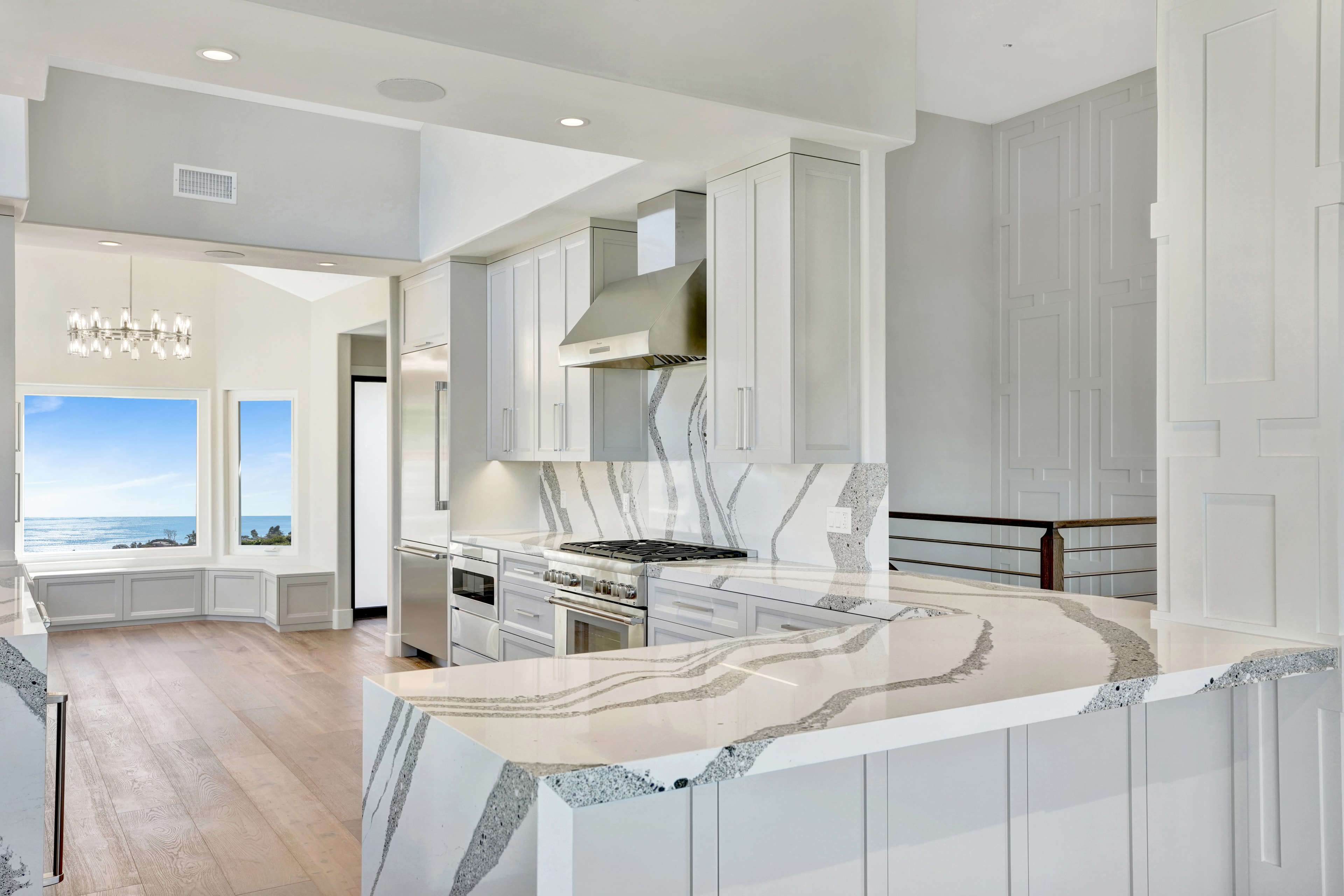 Kitchen remodel in Laguna Beach CA — white shaker upper and lower cabinets with book-matched quartz slab backsplash behind professional range, stainless hood, and chandelier with ocean view through bay window by Main Street Kitchen & Flooring