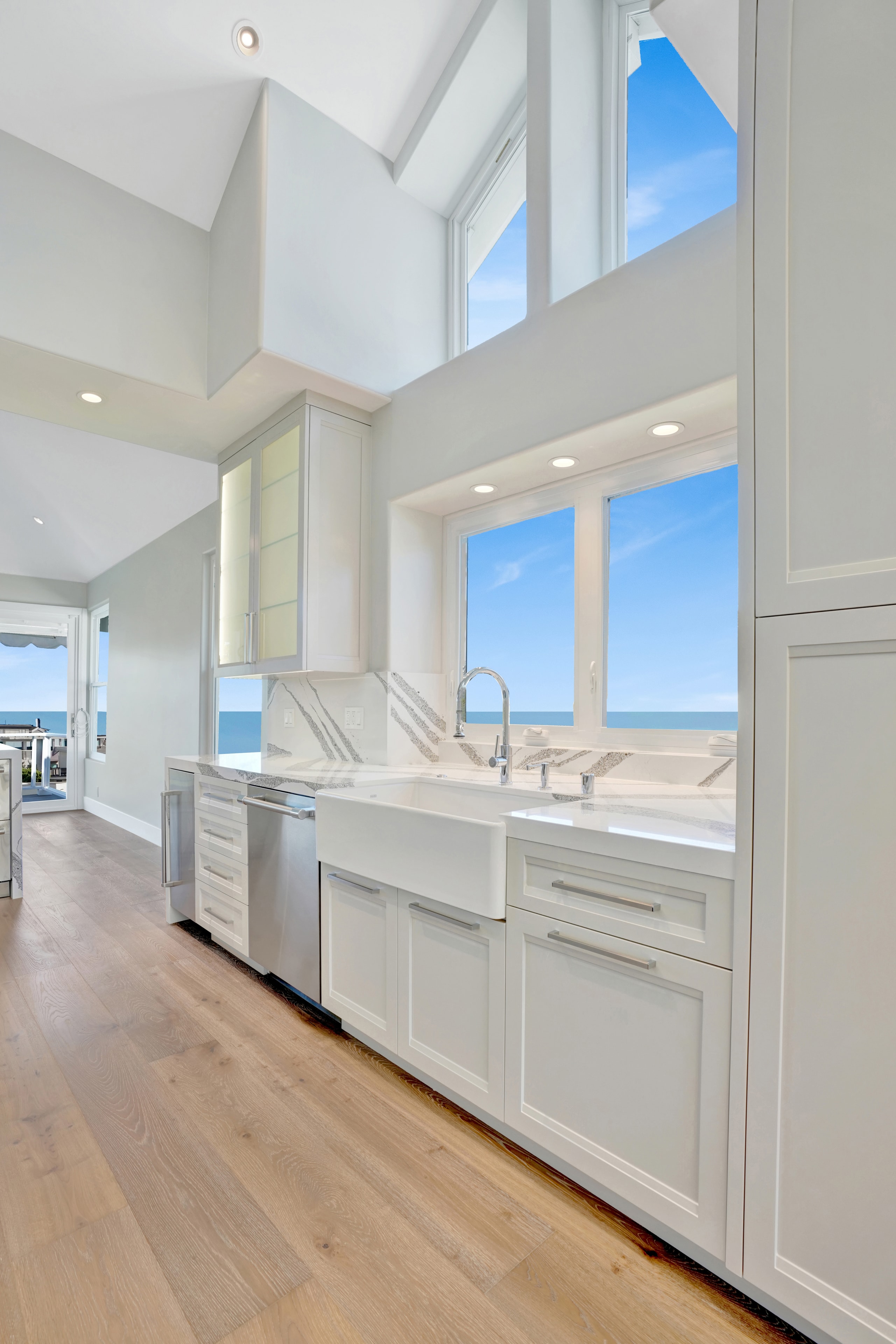 Kitchen remodel in Laguna Beach CA — white shaker cabinets with professional gas range, book-matched quartz slab backsplash, stainless hood, and ocean view through window at angle by Main Street Kitchen & Flooring