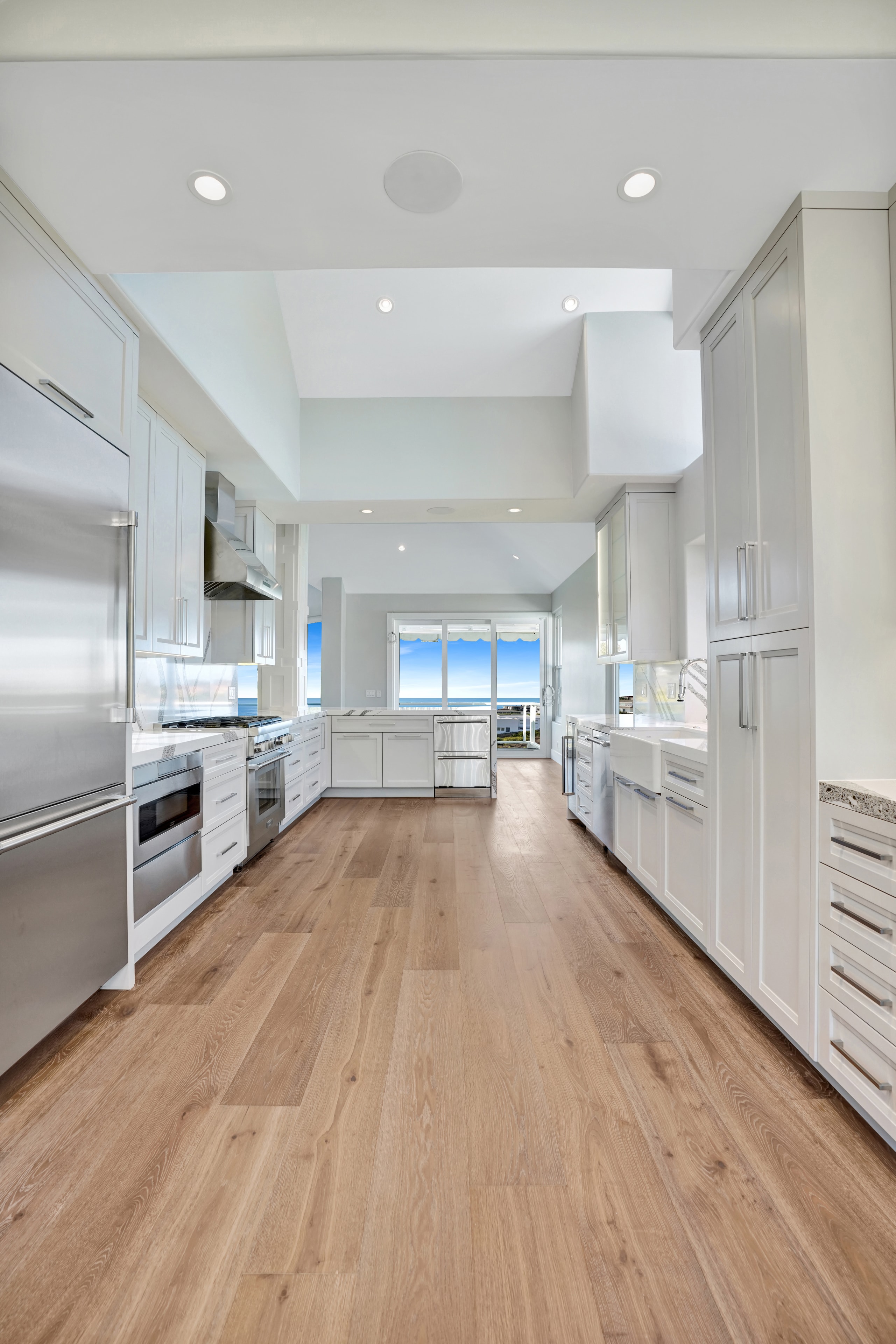 Kitchen remodel in Laguna Beach CA — white shaker cabinetry with vaulted ceiling, tall clerestory windows, farmhouse apron sink, chrome faucet, quartz backsplash, and panoramic ocean view by Main Street Kitchen & Flooring