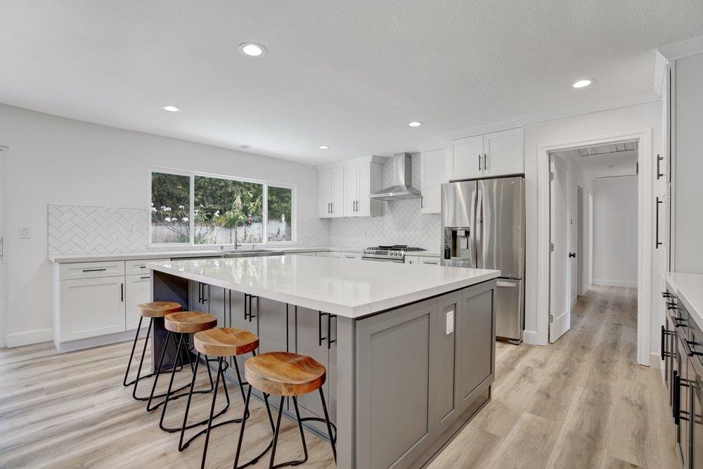 Open-concept kitchen remodel in Tustin CA — large gray island with quartz countertop and wood bar stools, white shaker cabinets, herringbone backsplash, stainless refrigerator, and light wood-look vinyl plank flooring by Main Street Kitchen & Flooring