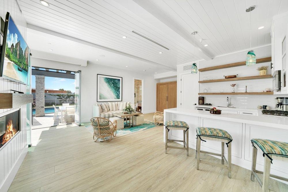 Living room remodel in Laguna Beach CA — coastal open-concept layout with shiplap wood beam ceiling, linear fireplace, light luxury vinyl plank flooring, open shelving, and bi-fold glass doors opening to pool patio by Main Street Kitchen & Flooring