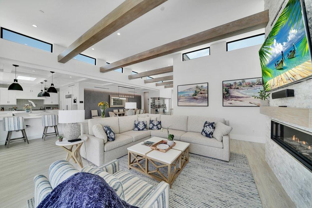 Living room remodel in Laguna Beach CA — grand open-plan living with exposed wood beam ceiling, skylights, stone fireplace surround, light wide-plank flooring, and open kitchen with white cabinets and black pendants by Main Street Kitchen & Flooring