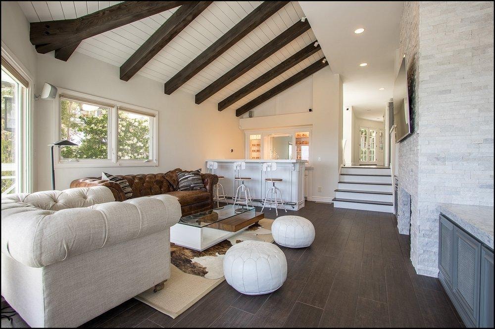 Living room remodel in Laguna Beach CA — vaulted ceiling with dark wood beams, stacked stone fireplace surround, dark hardwood flooring, wet bar with bar stools, and abundant natural light by Main Street Kitchen & Flooring