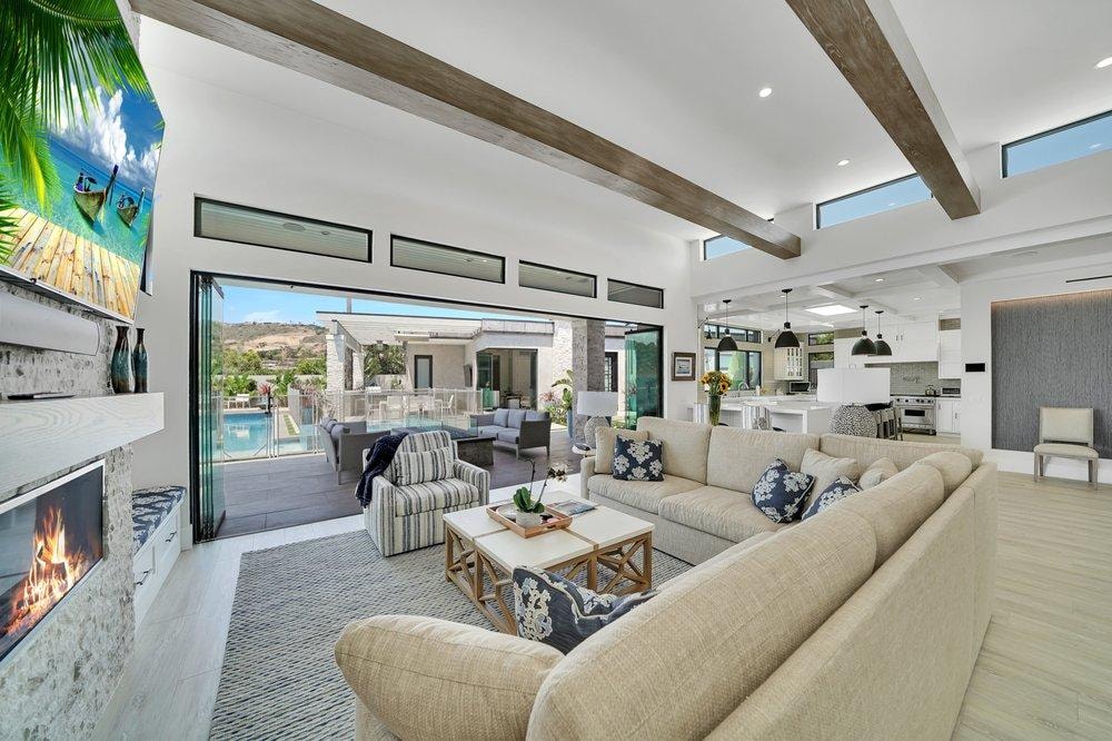 Living room remodel in Laguna Niguel CA — open concept layout with exposed wood beam ceiling, stone fireplace, light wood-look vinyl plank flooring, bi-fold glass doors opening to pool and outdoor living area by Main Street Kitchen & Flooring