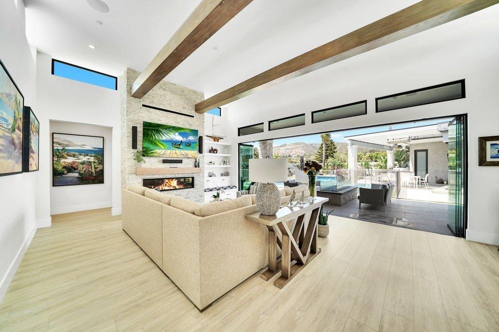 Living room remodel in Laguna Niguel CA — vaulted ceiling with wood beams, floor-to-ceiling stone fireplace surround, built-in shelving, light wide-plank wood-look flooring, and bi-fold glass doors with hillside views by Main Street Kitchen & Flooring